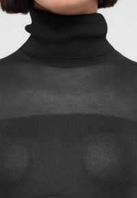 Γυναίκα που φοράει μαύρο ημιδιαφανές πλεκτό πουλόβερ με ρόμβο, εστίαση στην περιοχή του λαιμού και του άνω στήθους.