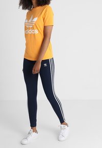 T-shirt en coton jaune avec le logo blanc d'Adidas, associé à des leggings navy avec trois bandes blanches sur les côtés, et des baskets blanches.