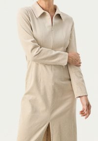 Robe en velours côtelé beige avec fermeture éclair à l'avant et col. Manches longues et fente latérale ajoutent du détail au design. Tissu doux et texturé.