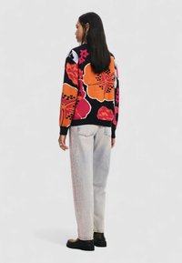Maglione lavorato a maglia con grandi e vivaci motivi floreali arancioni e rosa su sfondo nero, abbinato a jeans di denim chiaro e stivali neri.