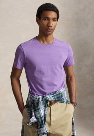Joven con el pelo corto y rizado, lleva una camiseta morada y una camisa verde a cuadros anudada alrededor de su cintura, con pantalones beige y las manos en los bolsillos.