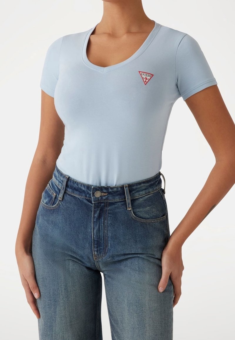 T-shirt ajusté bleu clair avec un col en V et des manches courtes, arborant un logo triangle rouge sur la poitrine. Associé à un jean en denim taille haute.