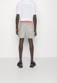 Pantalones cortos grises de tejido texturizado, con un bolsillo de parche. Llevados con una camisa blanca de manga corta y sandalias negras.