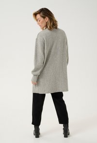 Grauer, übergroßer Pullover mit langen Ärmeln, gerippten Bündchen und einer weichen Textur. Kombiniert mit schwarzen Hosen und Stiefeletten.