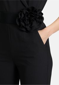 Czarny kombinezon z szerokim teksturowanym paskiem ozdobionym dużym, warstwowym kwiatem z czarnej tkaniny, z widocznymi kieszeniami po bokach.