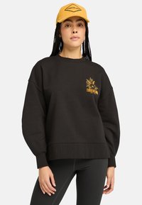 Schwarzer Sweatshirt mit lockerer Passform, gerippten Bündchen und goldfarbenem Timberland-Logo. Trägt es mit schwarzen Leggings und einer senfgelben Kappe.