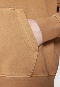 Main d'une personne partiellement dans la poche avant d'un sweat-shirt couleur beige avec couture visible et bord-côte au poignet.