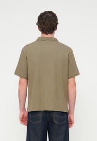 Πολύχρωμο πόλο πουκάμισο σε χακί χρώμα με υφασμάτινο ριμπ σχέδιο, κοντά μανίκια και κλασικό γιακά. Η πίσω όψη δείχνει χαλαρή εφαρμογή και διακριτικά πλαϊνά ανοίγματα.