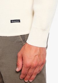 Kremowy sweter dziany z prążkowanymi mankietami, z czarną etykietą "ArmorLux" przy dole, w połączeniu z teksturowanymi brązowymi spodniami.
