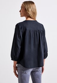 Blusa nera a maniche lunghe con scollatura rotonda, dettaglio arricciato sulla schiena e polsini con bottone, abbinata a jeans grigi chiari. Texture del tessuto liscia.