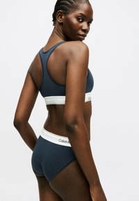 Femme aux cheveux tressés portant un soutien-gorge de sport Calvin Klein bleu marine et des sous-vêtements assortis, posant de dos et de profil sur un fond blanc.