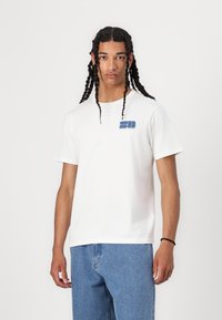 Vit bomullströja med korta ärmar, har ett blått grafiskt emblem på vänster bröstområde. Rakt skuren design kombinerad med blå denimshorts.