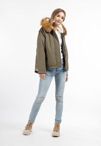 Olivengrøn jakke med en kunstpels hætte, lynlås-lukning og en blød, foret inderside. Revne lyseblå jeans og beige høj-top sko.