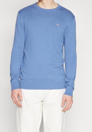 Man draagt een lichtblauwe trui met lange mouwen en een klein geborduurd logo op de borst, gecombineerd met een witte broek, tegen een witte achtergrond.