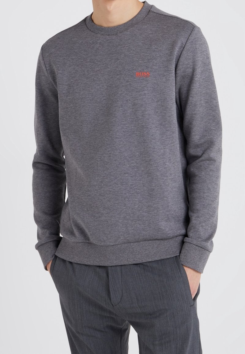 Homme portant un sweat-shirt gris à col rond avec un petit logo rouge « BOSS » sur le côté gauche de la poitrine, et un pantalon gris foncé, les mains dans les poches.