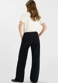 Pantalones de pana negra de corte holgado, con bolsillos traseros y una superficie texturada. Llevados con una camiseta blanca desde un ángulo trasero.