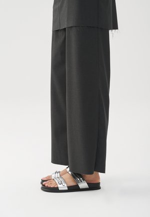 Melni bikses ar brīvu piegriezumu un neapstrādātu apdari, pārī ar plakano kurpju sandālēm, kas aprīkotas ar sudraba siksnām un dekoratīvām detaļām.