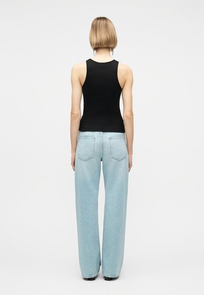 Άτομο με κοντά ξανθά μαλλιά που φοράει μαύρη αμάνικη μπλούζα και ανοιχτό μπλε παντελόνι με φαρδιά μπατζάκια, στέκεται γυρισμένο προς τα πίσω σε απλό φόντο.