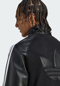 Czarna sportowa kurtka z połączeniem gładkiej skóry i tekstury siatkowej, z białymi pasekami i wyraźnym czarnym logo Adidas na plecach.