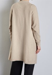 Béžový pletený cardigan s dlouhými rukávy, žebrovanými manžetami a volným střihem, s rozparkem na spodním lemu a texturovanými detaily na ramenou.