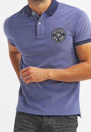 Mann trägt ein blaues Polohemd mit dunklem Kragen und einem runden Hilfiger New York Logo-Patch auf der linken Brust, kombiniert mit dunkler Jeans.