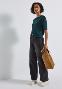 Haut teal à manches courtes avec une texture lisse, associé à un jean noir taille haute. Tient un sac en daim beige ; porte des baskets blanches avec des semelles crème.