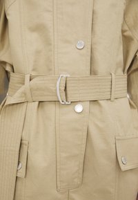 Veste en coton beige avec une taille cintrée, dotée d'une ceinture à boucle plate, de boutons-pression et de deux poches avant. Tissu texturé avec des détails de couture.