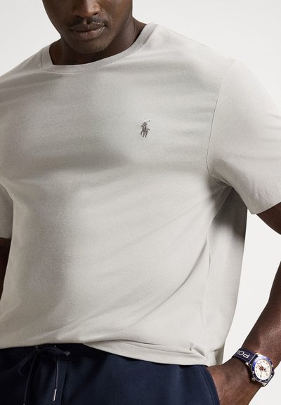 Ανοιχτό γκρι κοντομάνικη μπλούζα από μαλακό ύφασμα, με μικρό κεντημένο λογότυπο στο στήθος, φορεμένη με σκούρο μπλε παντελόνι και ρολόι.