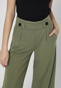 Žena oblečená v olivově zelených kalhotách s vysokým pasem a černými knoflíky, jedna ruka v kapse a druhá spuštěná podél těla.