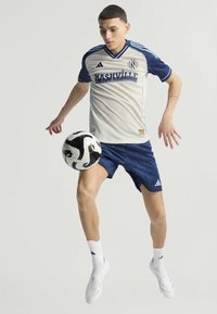 Maillot Adidas gris clair avec des accents bleu marine, comportant le texte "Nashville". Associé à un short bleu marine et des chaussettes blanches, avec un ballon de football noir et blanc.