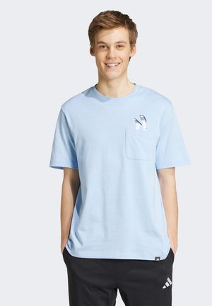 Młody mężczyzna uśmiecha się, ubrany w jasnoniebieską koszulkę z małym nadrukiem stóp w sandałach na kieszeni oraz czarne spodnie Adidas.