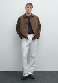 Bruine knoopjack met een grijze kraag, zwart shirt eronder, witte broek en zwarte schoenen. Casual fit, gladde stof en duidelijke zakken.