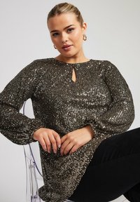 Top in paillettes nere e oro luccicanti con scollatura a chiave e maniche lunghe a sbuffo. Tessuto morbido, vestibilità casual, indossato con pantaloni neri.