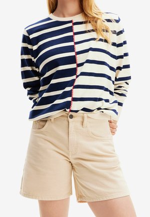 Vrouw die een marineblauw-wit gestreept shirt met lange mouwen draagt, met een rode zigzagnaad en beige shorts met hoge taille.