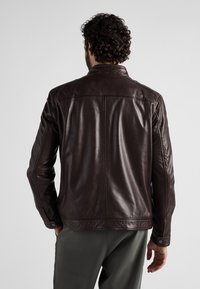 Brun läderjacka med en slät yta, utrustad med en spetsig krage, knäppta ärmslut och en klassisk passform. Bärs över grå byxor.