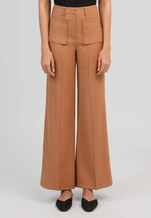 Pantalon marron à jambes larges avec de grandes poches avant, porté avec des ballerines noires pointues et un haut noir, sur un fond gris clair.