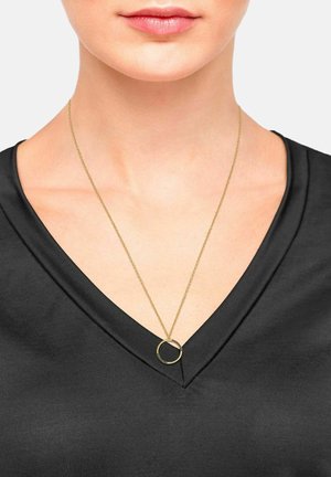 Collana d'oro con un pendente circolare, sospesa a una catena sottile, indossata sopra un top nero a V. Finitura in metallo liscia, design minimalista.