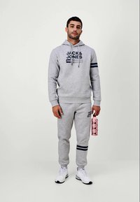 Sweat à capuche gris et pantalon de jogging en mélange de coton avec des accents marins, arborant un logo sur la poitrine et la jambe, associés à des baskets blanches et une bouteille d'eau à motifs.