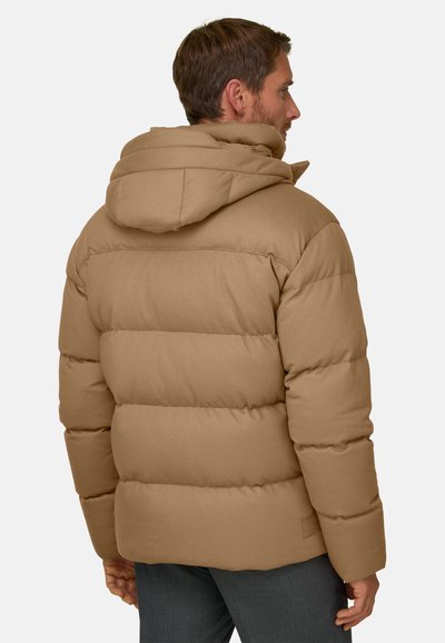 Homme portant une doudoune beige avec capuche, vu de dos et légèrement tourné sur le côté sur un fond uni.