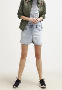 Un overol de mezclilla en azul claro con dobladillo enrollado, combinado con una camiseta gris y una chaqueta verde con detalles bordados. Zapatillas negras completan el look.