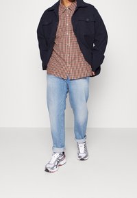 Chaqueta azul marino oscuro con dos bolsillos en el pecho, camisa de cuadros marrón, pantalones vaqueros de mezclilla azul claros y zapatillas atléticas plateadas con acentos rojos.