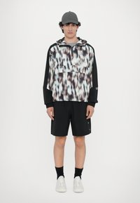 Pullover leggero e stampato con tasca frontale, maniche nere, abbinato a pantaloni corti neri e scarpe da ginnastica bianche. Un cappellino grigio completa il look.