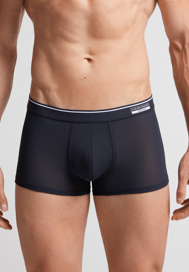 Mörkblå trunk-stil kalsonger med en mjuk, elastisk midjeband som har ett vitt streck och en logotypetikett. Slät, stretchig tyg, figurnära design.