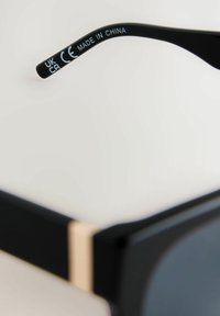 Lunettes de soleil noires avec une finition brillante, présentant un accent doré sur la branche. La branche comprend le texte : "UK CA CE MADE IN CHINA."