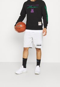 Czarna bluza z kapturem z fioletowym i zielonym napisem, szare szorty z logo "UNDER ARMOUR", czarne skarpetki z niebieskimi akcentami, trzymająca koszykówkę.