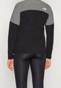 Persona che indossa una giacca a maniche lunghe nera e grigia della North Face e leggings neri lucidi, vista da dietro su uno sfondo semplice.