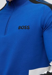 Primer plano de un suéter azul con cremallera, con rayas negras en los hombros y el logo "BOSS" bordado en el pecho.