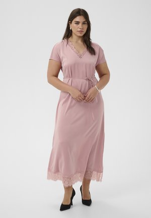 Femme portant une robe midi rose à bordure en dentelle, manches courtes, ceinture à la taille, boucles d'oreilles en perles, bracelet et escarpins noirs à bout pointu, debout sur un fond blanc.