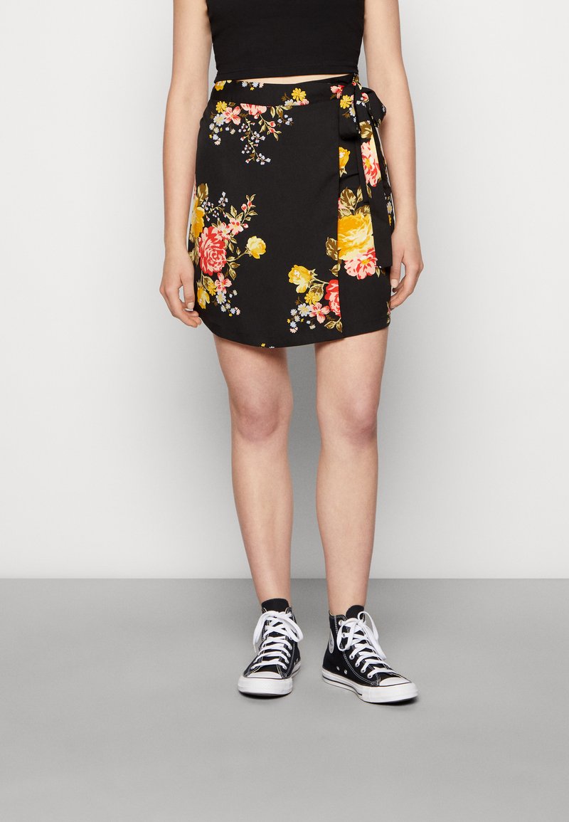 Saia envolvente preta com flores vibrantes amarelas e rosas, apresentando uma fita na cintura e uma bainha ligeiramente rodada. Usada com sapatilhas pretas.