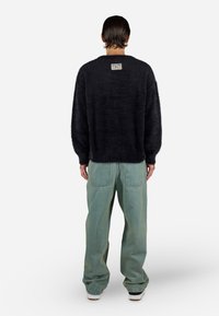 Sort fluffy sweater med lange ærmer, rund halsudskæring og et mærke på bagsiden, parret med lyseblå posede jeans og hvide sneakers.